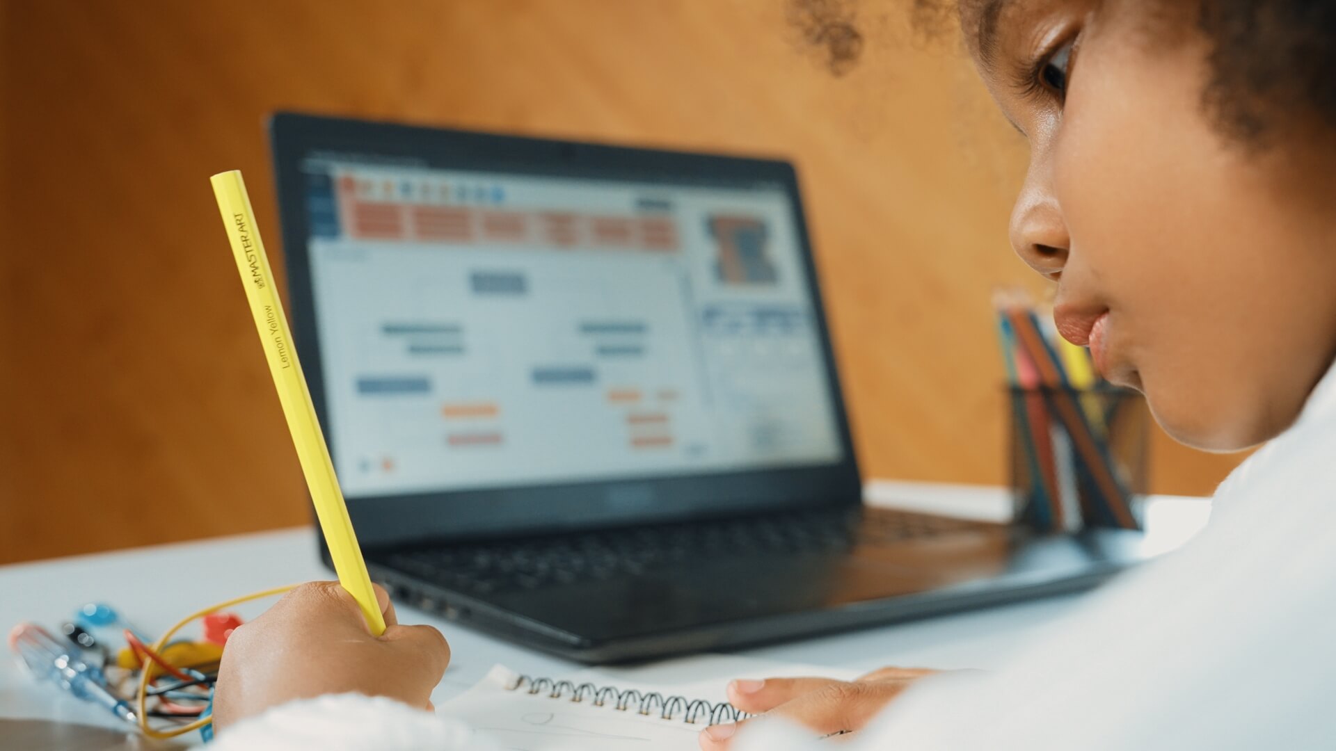 young Black girl writing in a notebook while online learning with a laptop